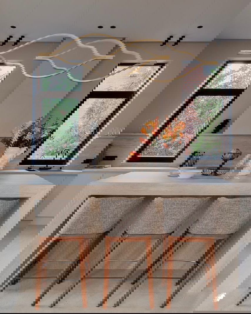 Кухонная зона с островом: барные стулья на деревянных ножках, две высокие рамы окон и волнообразная подвесная люстра над рабочей поверхностью.