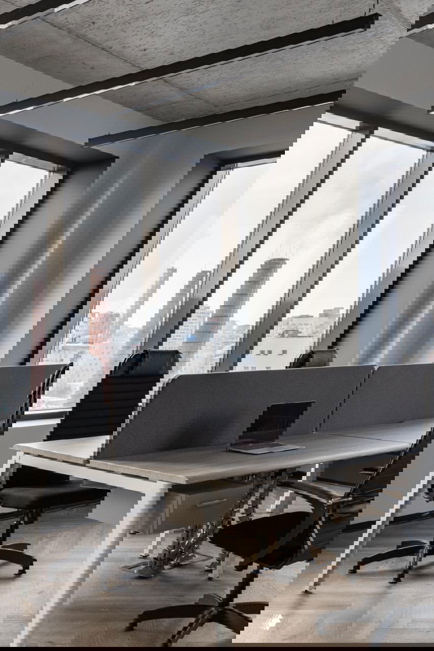 Общее рабочее пространство с большими окнами, офисными столами и креслами, вид на городской пейзаж через панорамные окна.