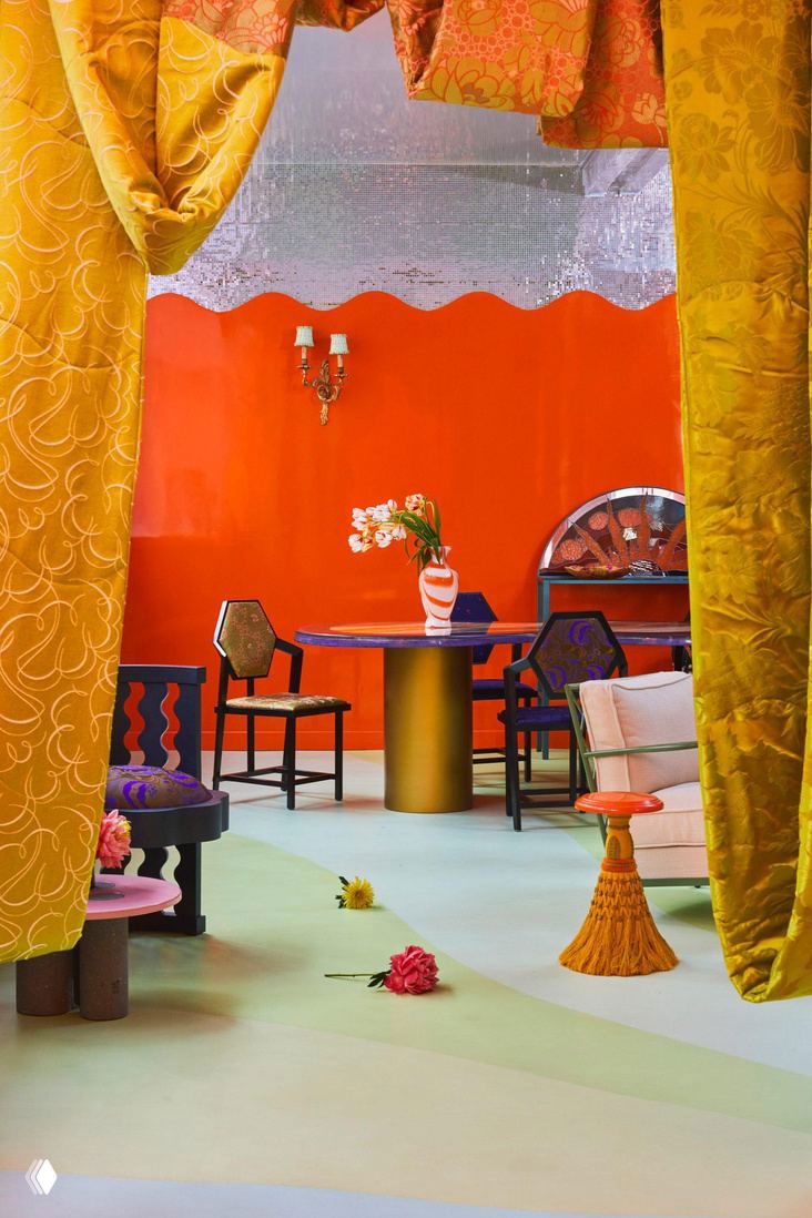 Интерьер с тяжелыми золотистыми драпировками, оранжевой стеной и столом с золотыми опорами — сочетание барочного и футуристического языков дизайна.