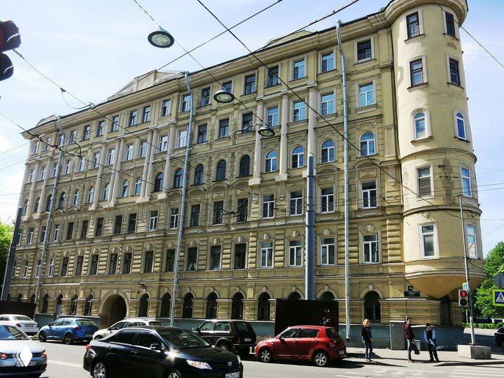 Фасад жилого дома в Петербурге: многоэтажный дом с арочными входами и балконами, припаркованные автомобили у тротуара.