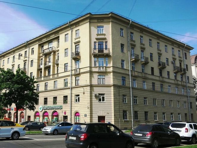 Угловой жилой дом в Санкт-Петербурге с магазинами в нижнем этаже, окна-эркеры и автомобили на перекрёстке возле фасада.