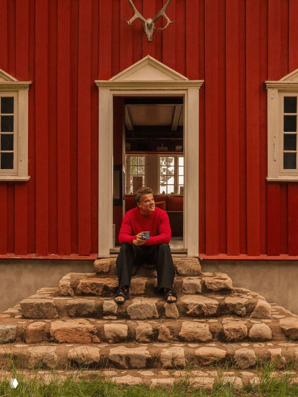 Бьярке Ингельс сидит на каменных ступенях у входа в красный коттедж, держит кружку, на заднем плане видно внутреннее помещение и окно с видом на сад.