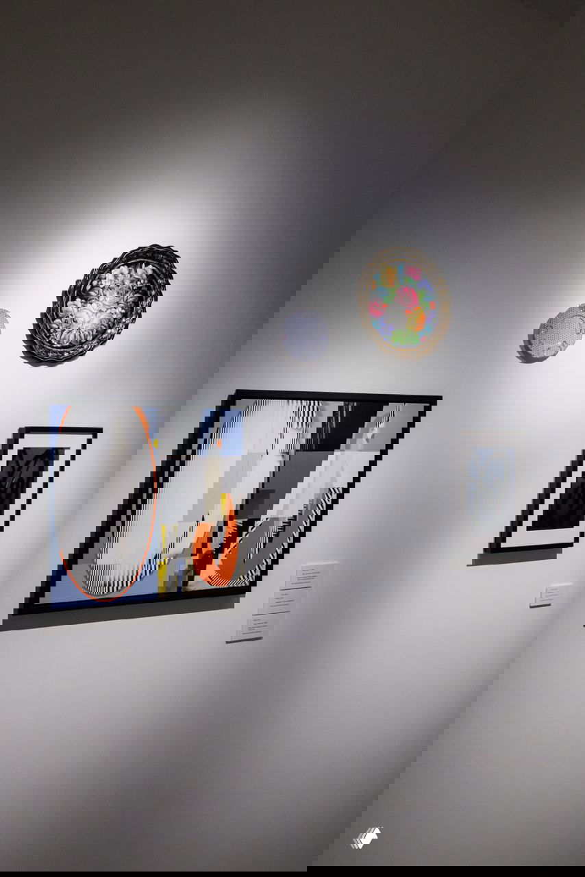 Стена галерейного зала с тремя оформленными работами: крупная картина, декоративная тарелка и рамочная графика под точечным светом.