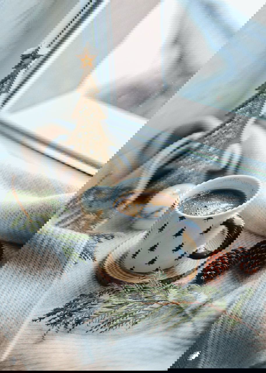 Кружка с горячим кофе на подставке у окна рядом с маленькой декоративной елочкой, шишками и текстилем — уютный зимний уголок для передышки во время подготовки дома.