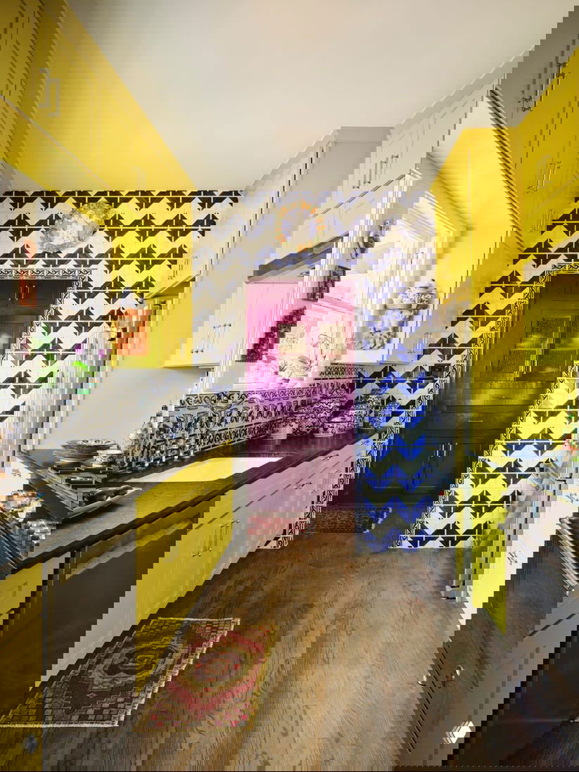 Кухня с ярко‑желтыми фасадами шкафов, узорной плиткой на стене и фиолетовой дверью в глубине коридора, сочетание смелых цветов и текстур.