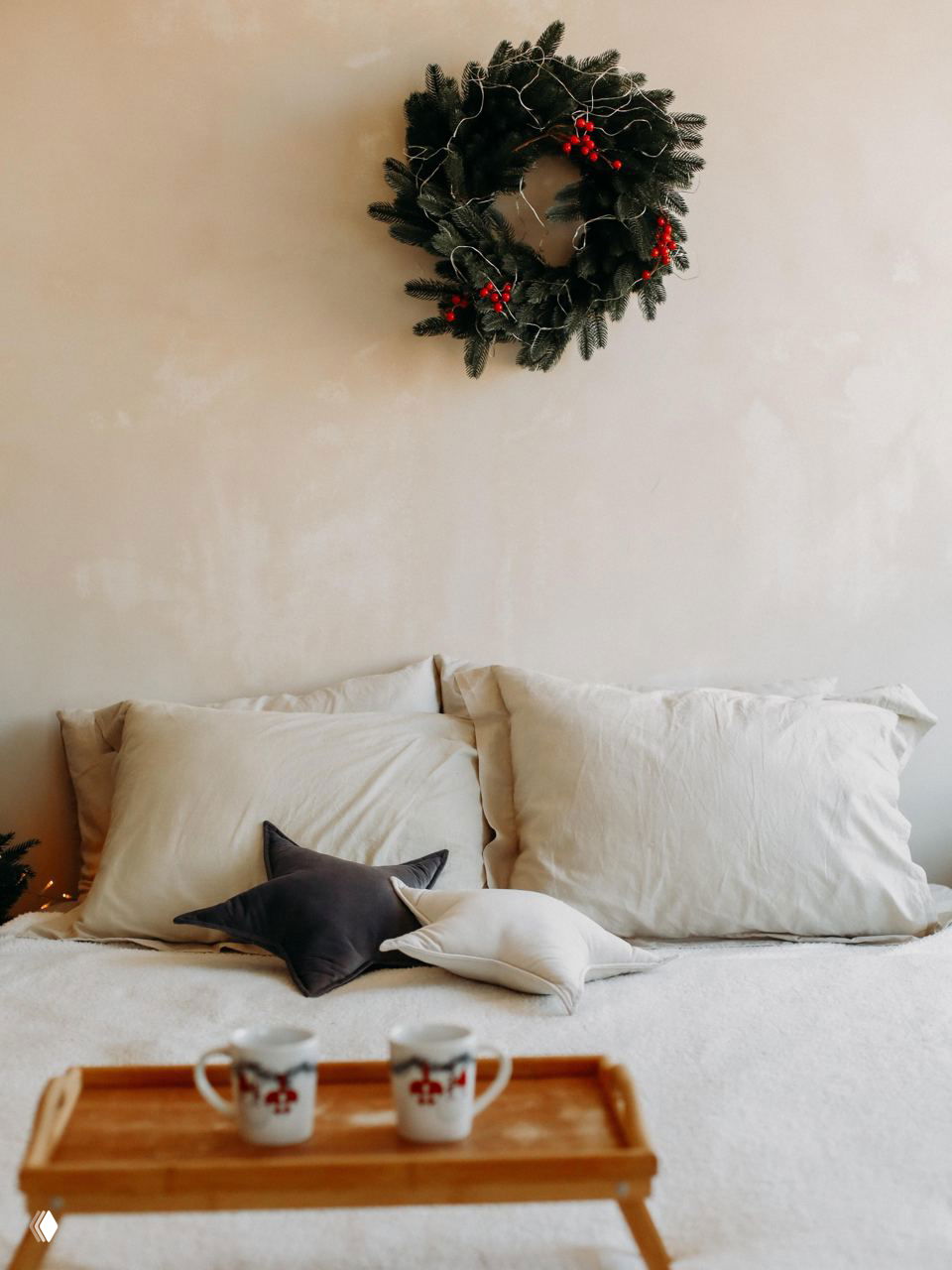 Рождественский венок на светлой стене над кроватью с подушками и подносом с чашками в минималистичной спальне — спокойный праздничный интерьер для отдыха.