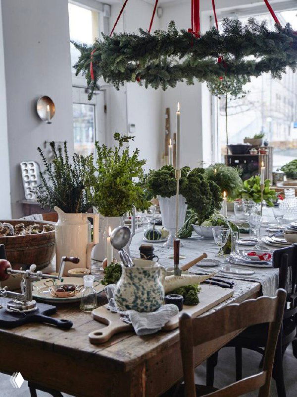 Круглая подвесная композиция из еловых ветвей над столом, свечи и зелёные акценты создают естественный зимний декор вместо привычной елки.