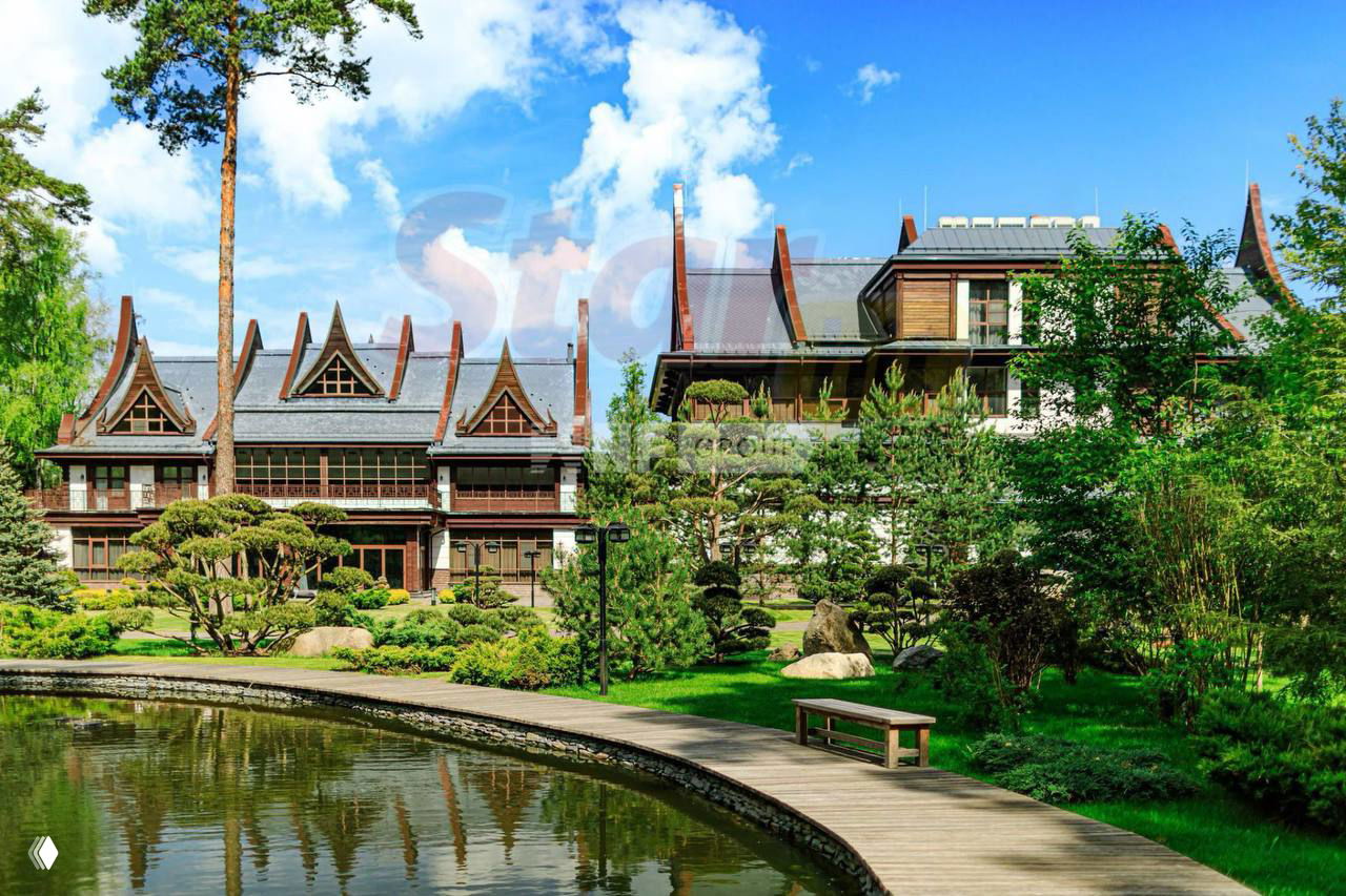 Внешний вид двух роскошных особняков на Новой Риге с озеленённой территорией, дорожками и декоративным водоёмом на фоне ясного неба.