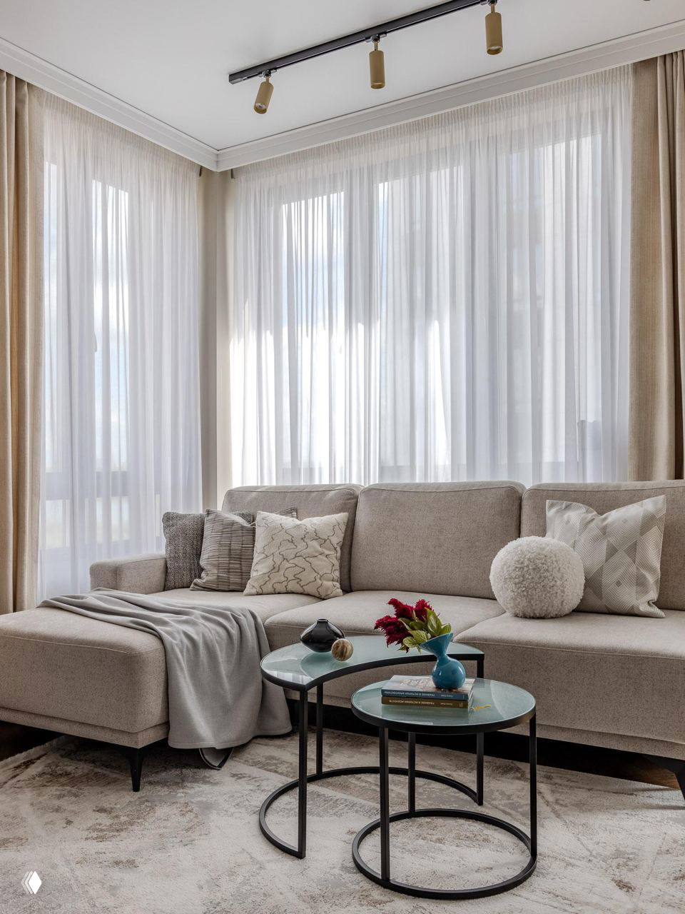 Гостиная с угловым бежевым диваном, круглыми стеклянными приставными столиками, светлыми шторами и декоративными подушками.