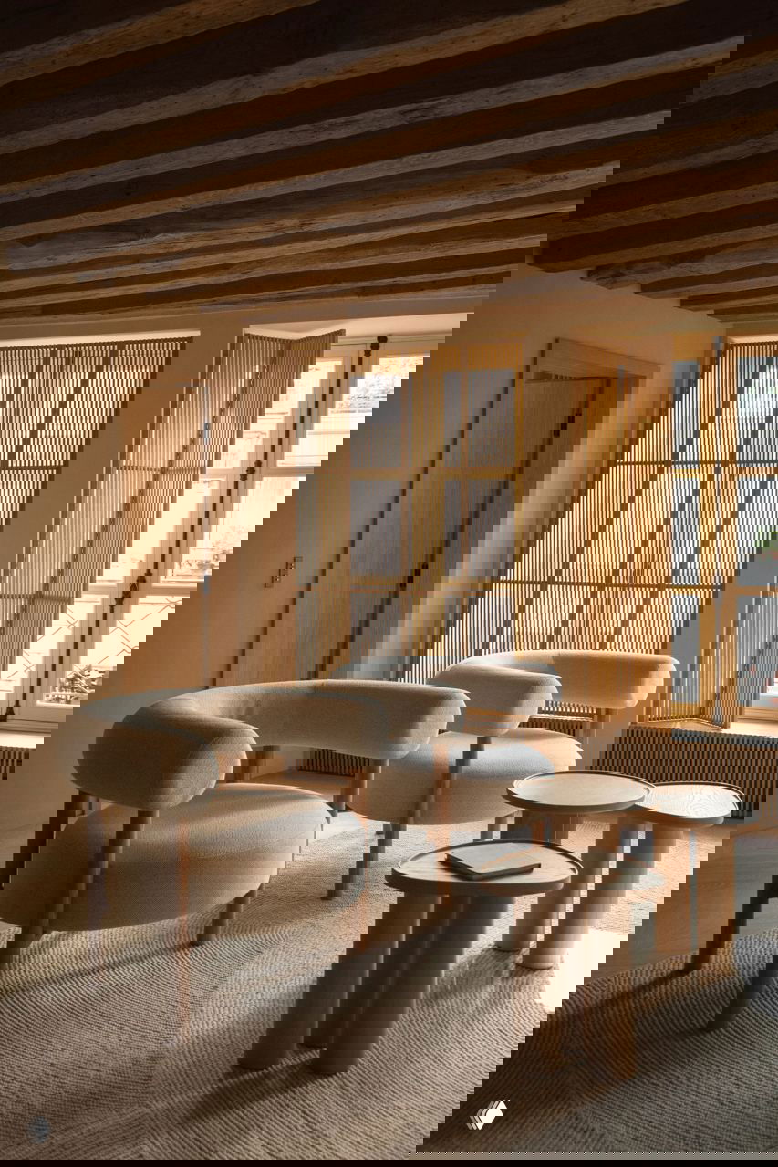 Интимная гостиная в парижской квартире с видимыми деревянными балками, светлыми округлыми креслами и вертикальными деревянными ставнями у окна.