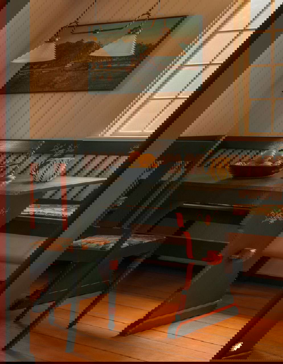 Обеденная зона с резным деревянным столом и скамьёй в тёмно-зелёных и красных тонах, окно пропускает мягкий свет, подчёркивающий натуральные текстуры интерьера.