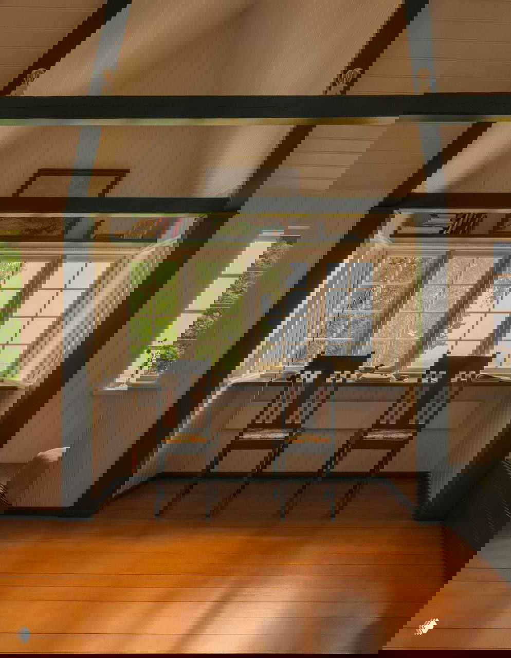 Рабочая зона у большого окна с двумя стульями и видом на лес; высокие балки и деревянный пол создают ощущение простора в мансарде летнего домика.