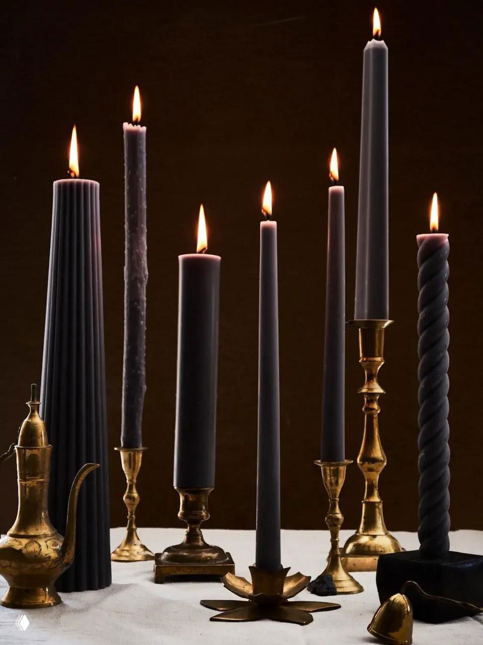 Несколько тонких и толстых черных свечей разных форм, зажженных и расставленных в латунных подсвечниках на темном фоне.