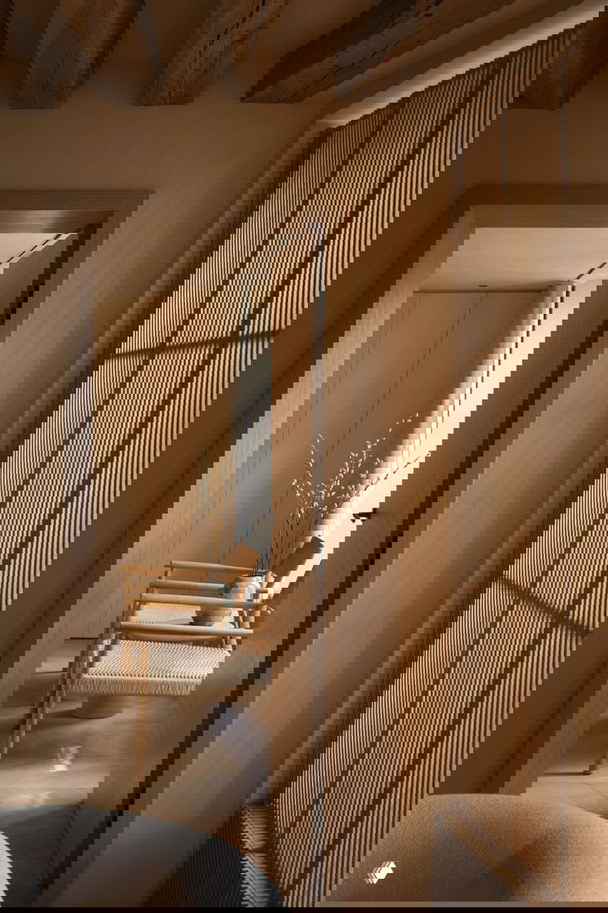 Вид через дверной проём на комнату с плетёным сиденьем, вазой с ветками и ровными древесными фактурами по периметру.