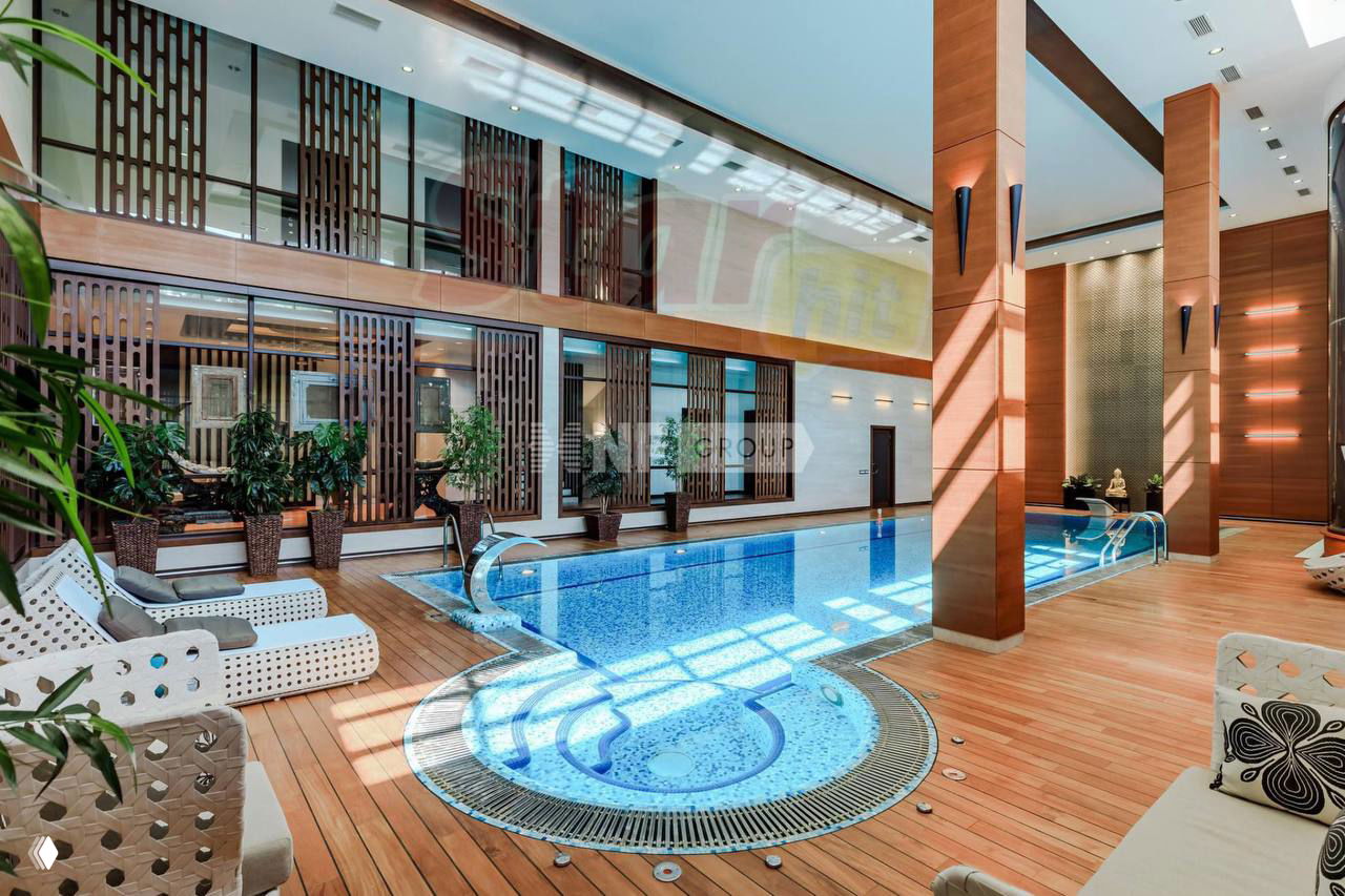 Крытый бассейн с зоной отдыха: большой стеклянный светлый зал, деревянный настил вокруг бассейна и шезлонги для комфортного времяпрепровождения.