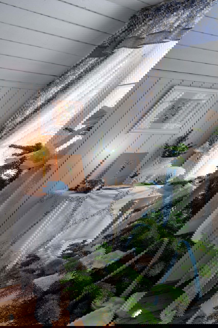 Уютный уголок спальни с украшенной ёлкой, кружевной скатертью на столе и мерцающей свечой — пастельная, романтичная новогодняя обстановка.