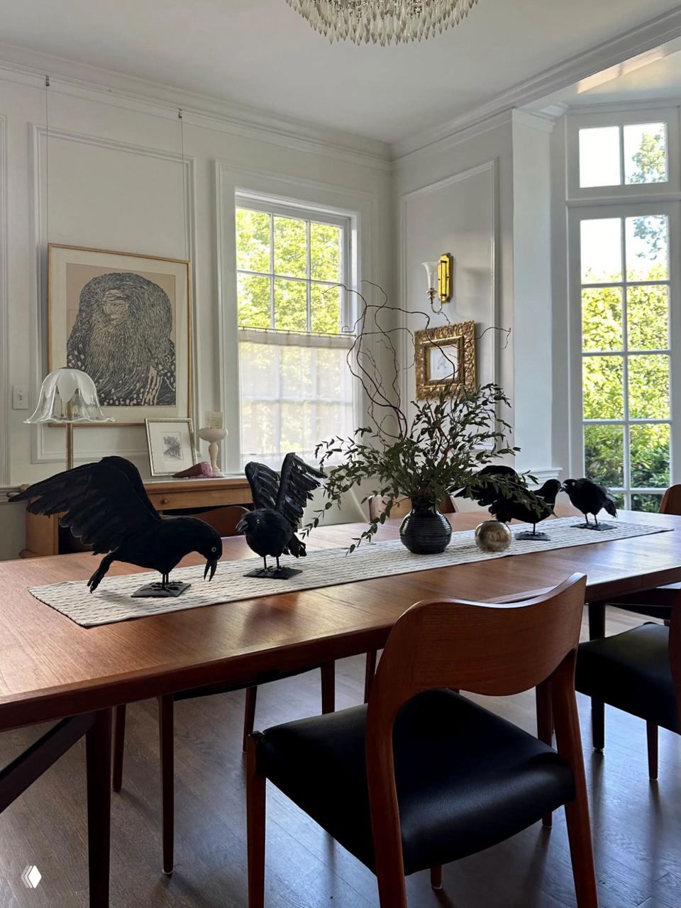 Обеденный стол с рядом черных декоративных воронов, зеленью в вазе и светлым интерьером с большими окнами на заднем плане.