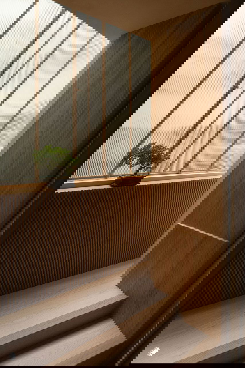 Ещё один ракурс лестницы у окна: мягкое освещение, тонкая деревянная отделка и одиночное кресло в светлой обивке.