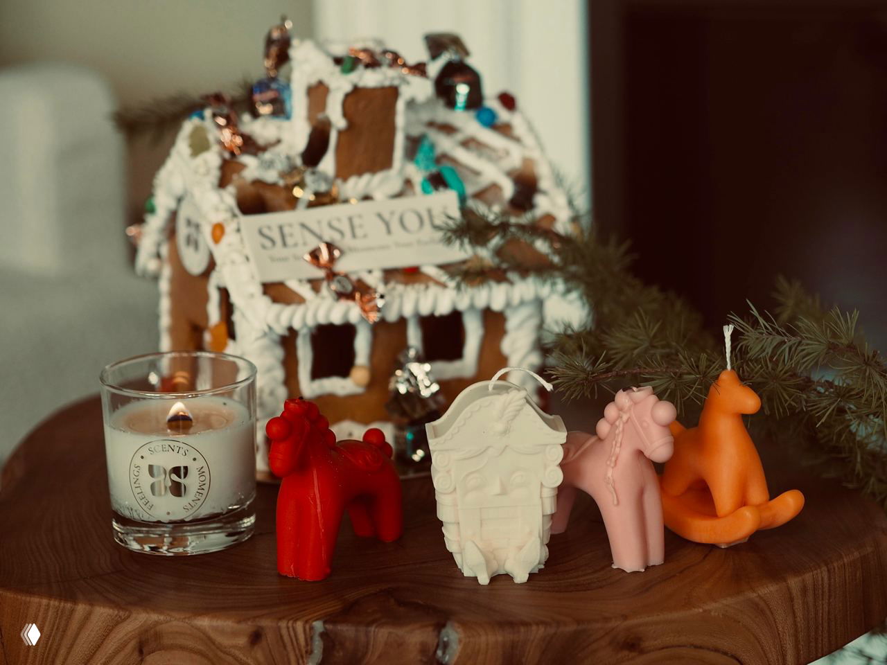 На деревянной поверхности пряничный домик и зажжённая свеча в стекле рядом с фигурными восковыми свечами‑лошадками в красном, белом и розовом цветах.