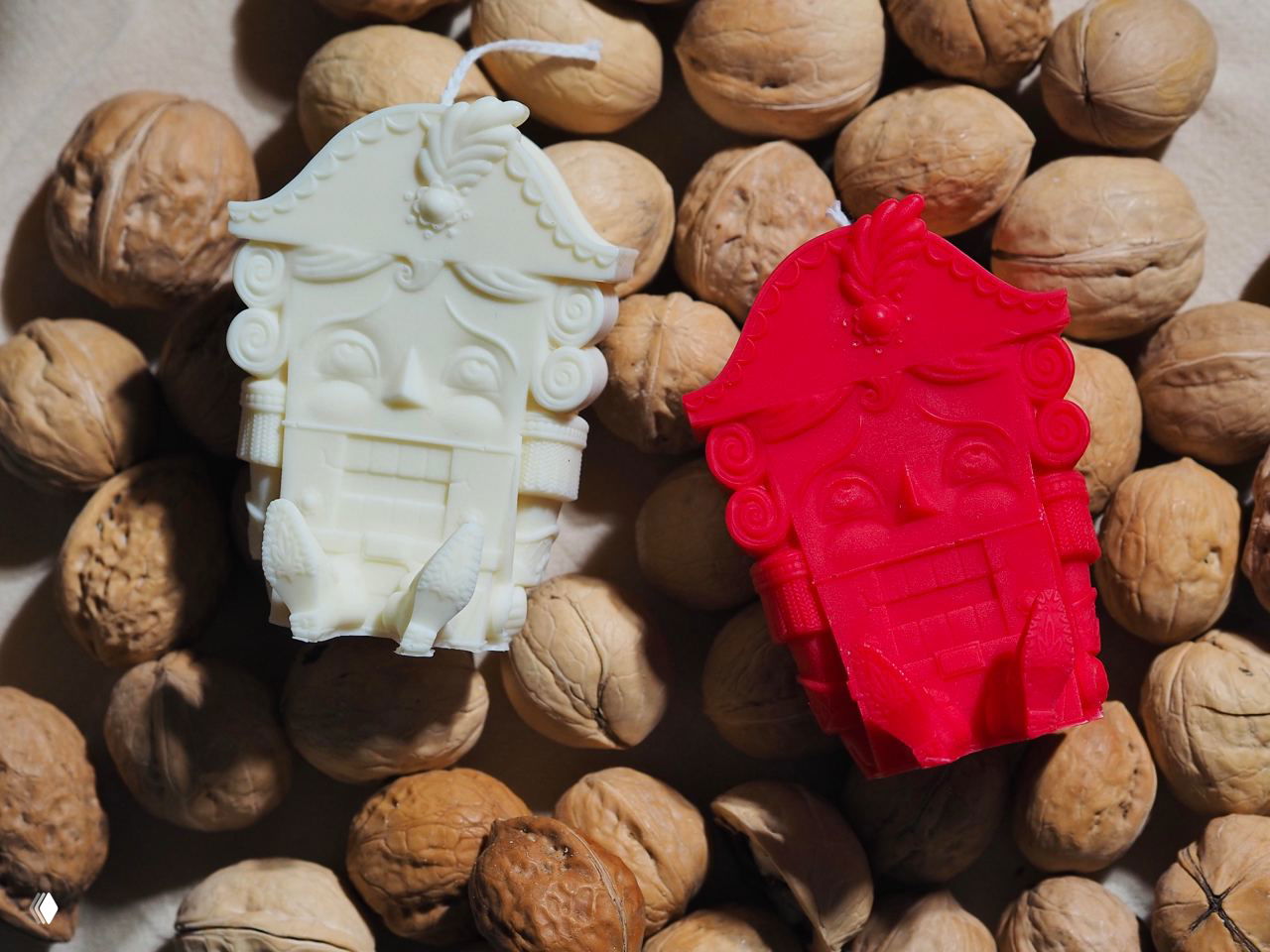 Две свечи‑Щелкунчика белого и красного цвета выложены на фоне скорлупы грецких орехов — детализированная форма с лицом и элементами декора.