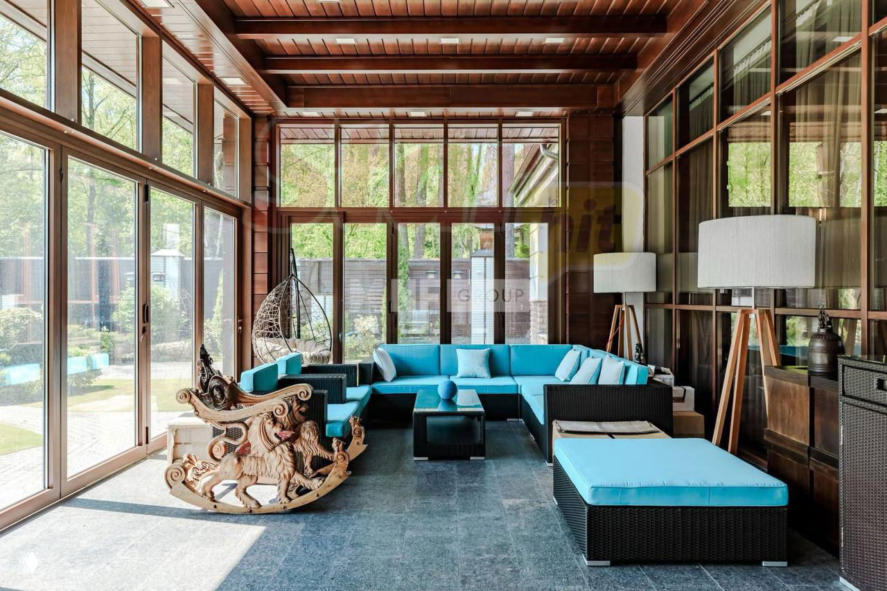 Зимний сад или веранда с яркими бирюзовыми мягкими модулями, деревянной отделкой и видом на внутренний двор частного участка особняка.
