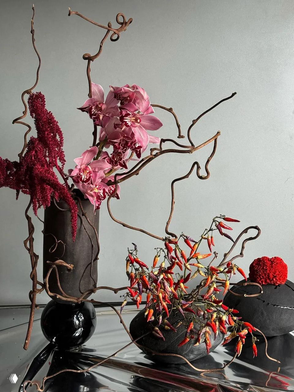 Высокая композиция из орхидей, веток и небольших ярких перцев чили в современных вазах на светлом фоне — драматичный флористический акцент.