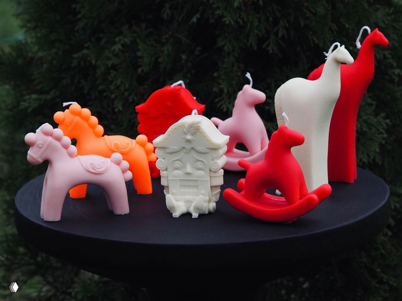 Групповой вид коллекции: несколько фигурных свечей — лошадки, Щелкунчик и высокий абстрактный силуэт, расположенные на чёрной подставке на фоне зелени.