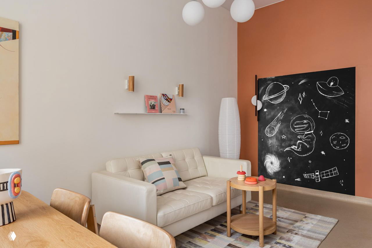 Уютный уголок ожидания с диваном, декоративной доской для рисунков и мягким ковриком; интерьер сочетает пастельные и тёплые оттенки для спокойной атмосферы.