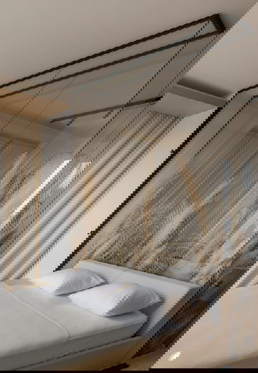 Спальня с прозрачной стеклянной перегородкой, за которой расположены высокие композиции сухоцветов, светлый текстиль и сдержанная нейтральная палитра.