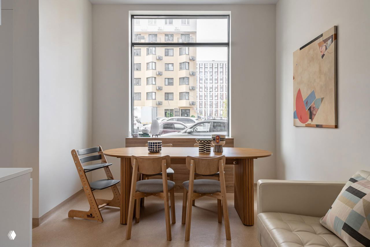 Столовая или переговорная зона клиники: овальный деревянный стол у большого окна, стулья из светлого дерева и элементы декора, которые делают пространство похожим на дом.