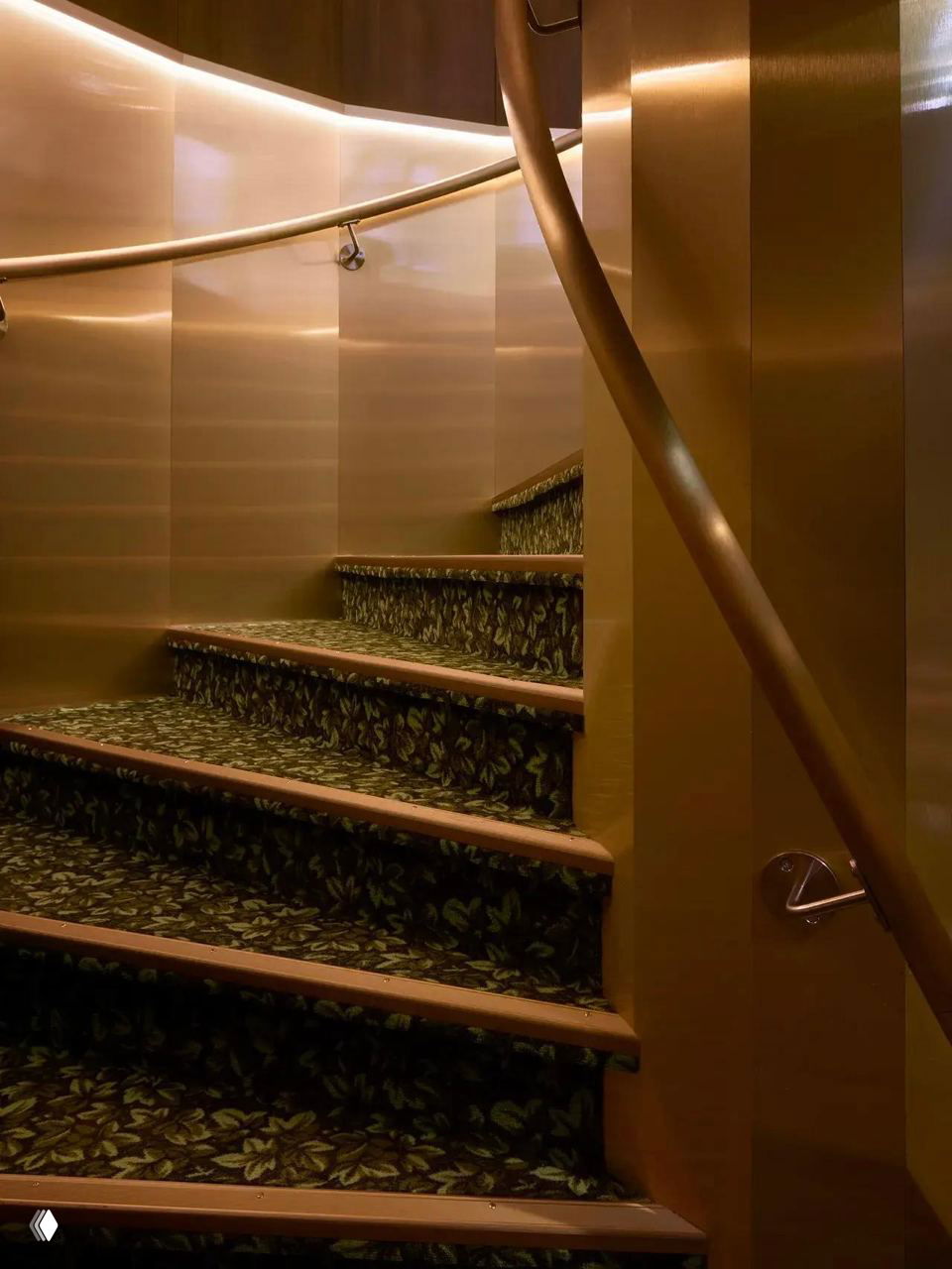 Золотистая винтовая лестница и перила в интерьере кинотеатра, детали отделки в теплых металлических тонах и узорчатый ковровый настил на ступенях.