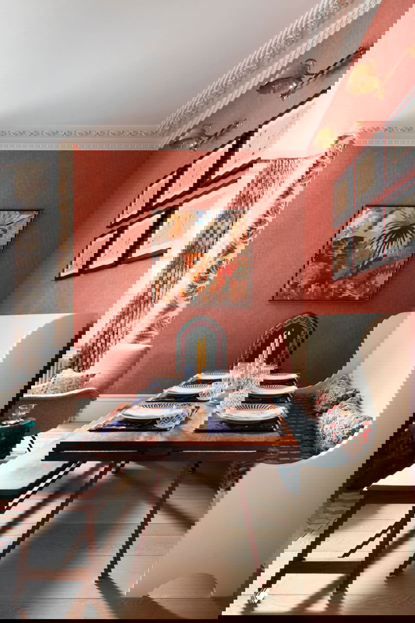 Столовая зона с деревянным столом, встроенной банкеткой с высокими спинками, красными акцентами и крупной картиной на фоне коралловых обоев.