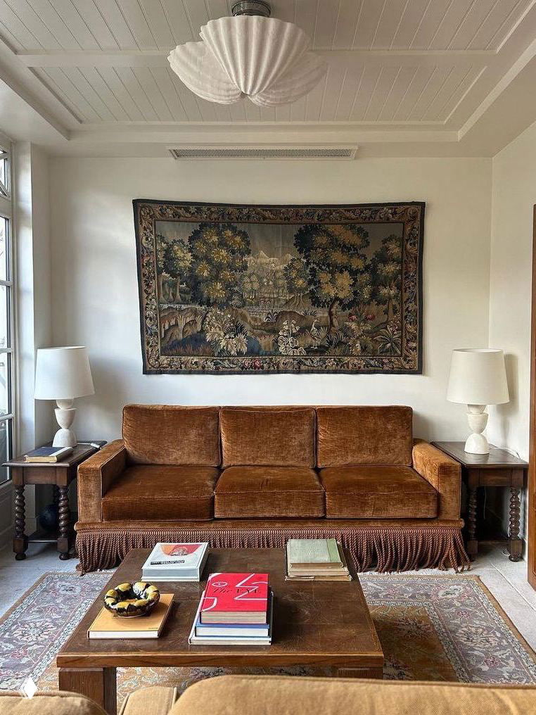 Коричневый бархатный трёхместный диван с густой бахромой по низу на фоне настенного гобелена и деревянных ночных столиков в уютной гостиной.