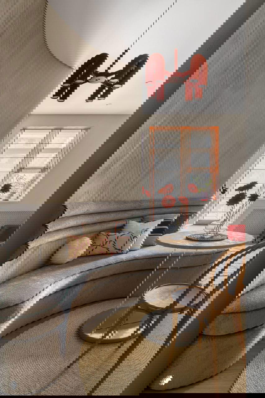 Гостиная с изогнутой фактурной стеной и полукруглым мягким диваном на фоне тёплого серого тона, заметны красные акценты и небольшой столик у окна.