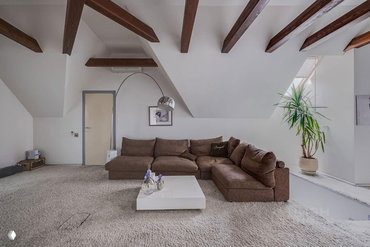 Гостиная на мансарде с коричневым угловым диваном, деревянными балками на потолке, низким белым журнальным столиком и комнатным растением у окна.