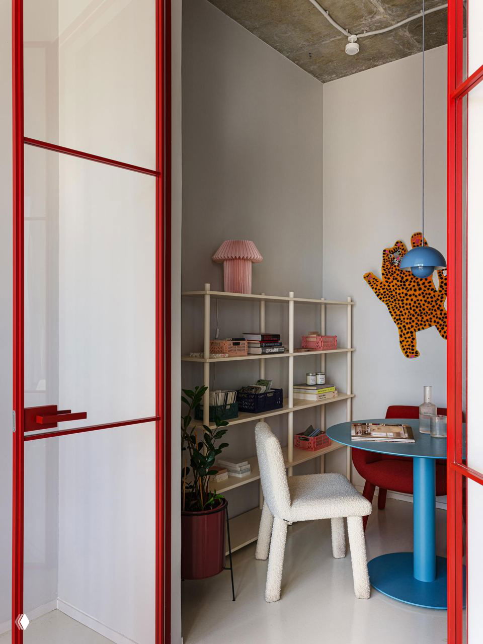 Небольшая рабочая ниша с красной рамой двери, круглым столом и уютным креслом; на стене декоративный элемент в виде пятнистой фигуры и полки с мелочами.