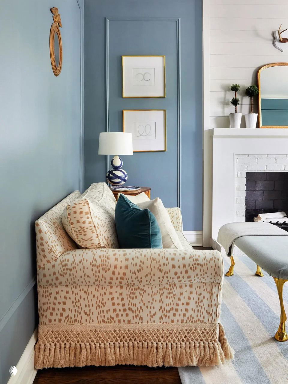 Светлый компактный диван с узорчатой обивкой и массивной декоративной бахромой в пастельной гостиной у камина; несколько подушек создают уют.