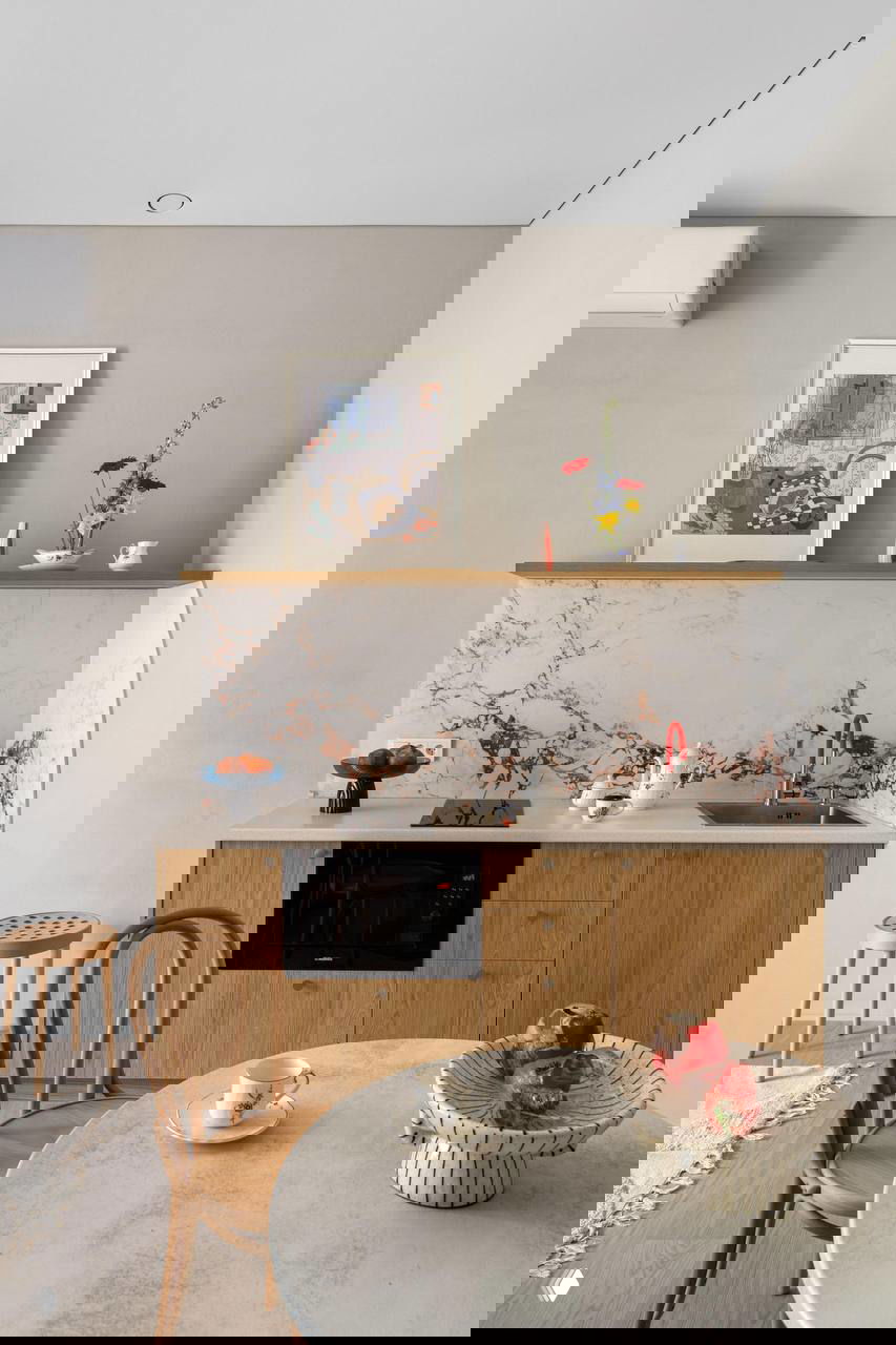 Кухонный уголок небольшой студии: деревянные фасады, мраморная столешница и яркий красный смеситель на нейтральном фоне стены.