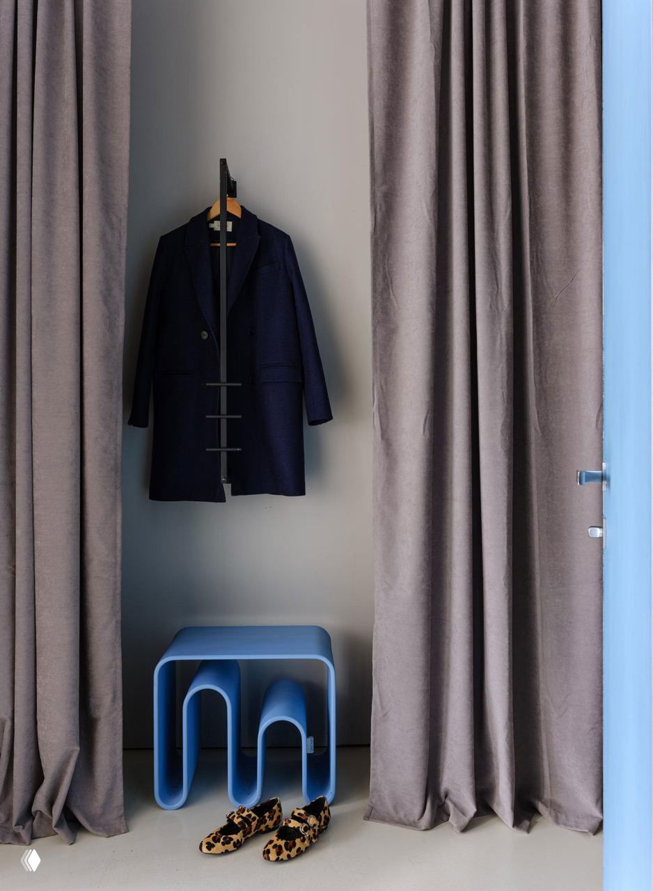 Уголок гардероба и хранения в офисе: вешалка с пальто, серые шторы и необычная синяя скамья-столик на фоне сдержанных стен студии DSGN HUB.