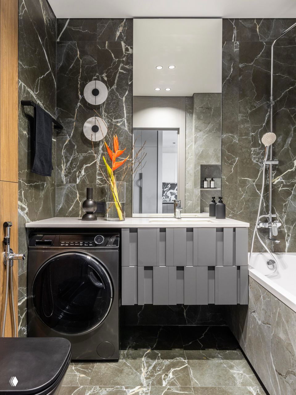 Ванная комната с тёмной мраморной облицовкой, стиральной машиной под раковиной, встроенной ванной и лаконичной минималистичной сантехникой.