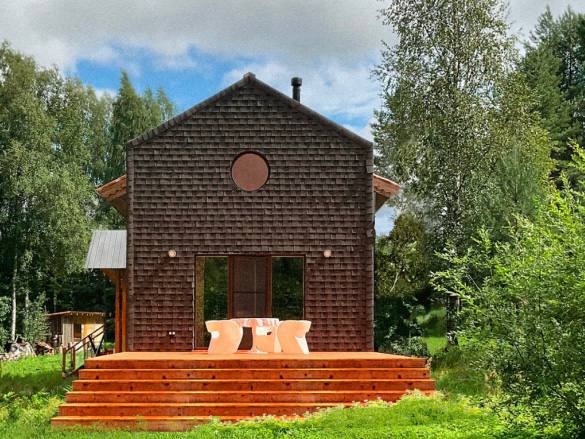 Тёмный деревянный дом с террасой и лестницей на фоне зелёного участка: акцент на текстуре дерева и простоте формы загородной архитектуры.