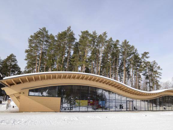 Современное волнообразное деревянное здание на заснеженной площадке: сочетание дерева, стекла и контраста с хвойным лесом на заднем плане.