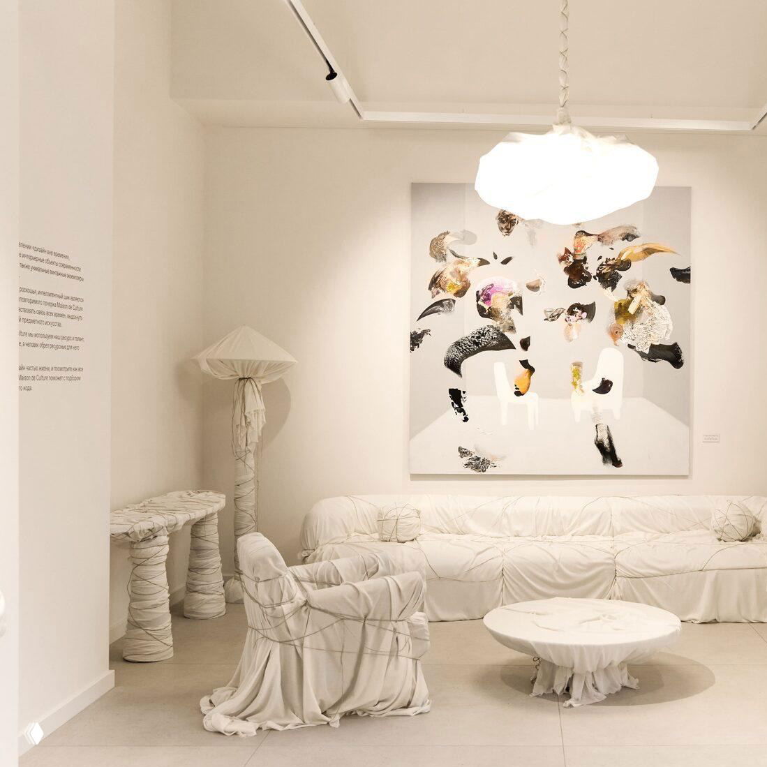 Светлый интерьер галереи с белой обтянутой тканью мебелью, низким круглым столом и большим абстрактным полотном на стене под мягким освещением