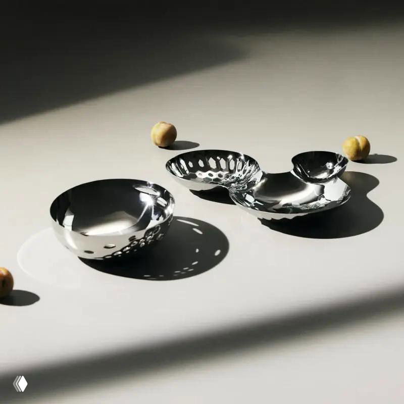 Несколько лаконичных стальных чаш и блюдец на световом фоне, игра света и тени на поверхности минималистичной коллекции посуды для сервировки.