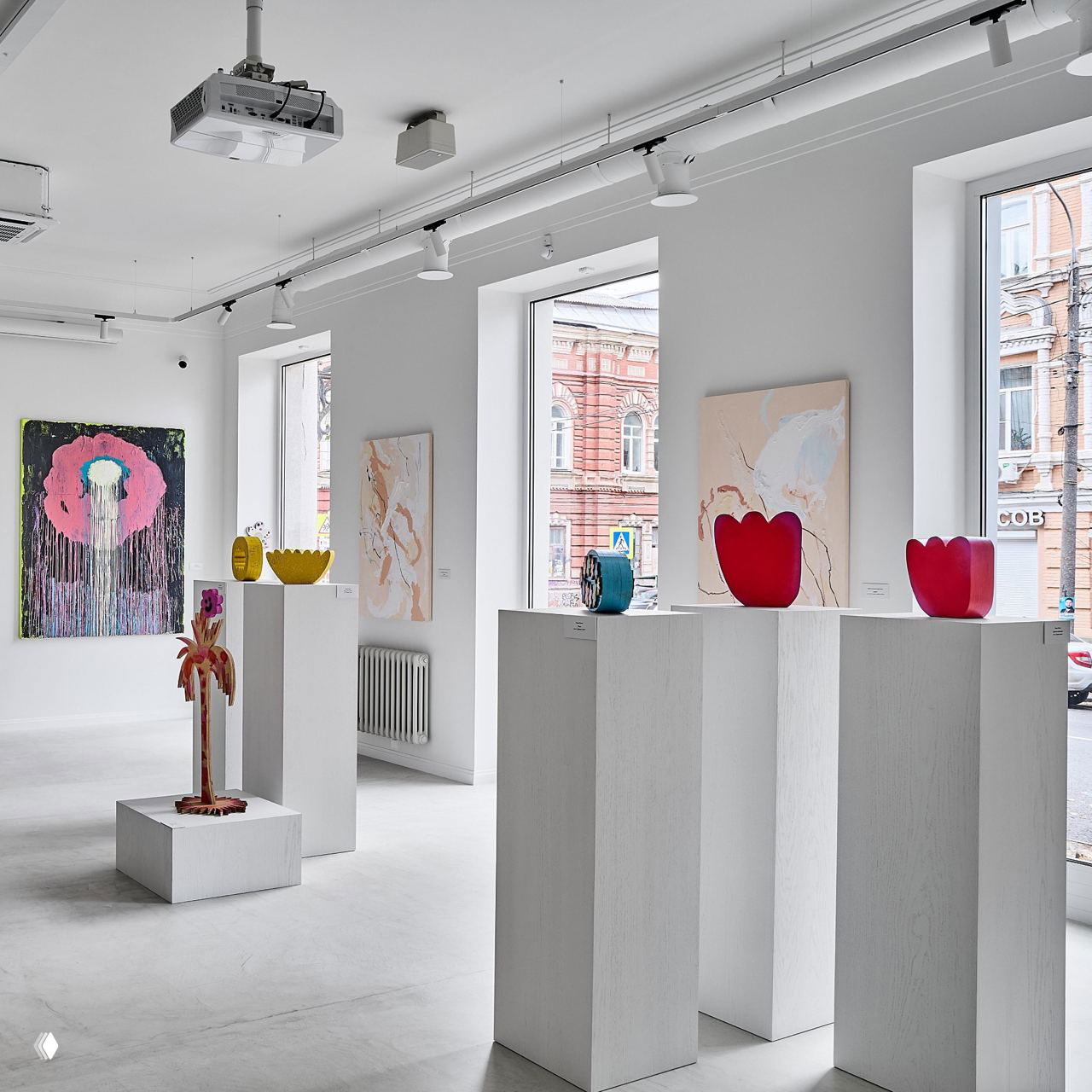 Яркая светлая экспозиция с белыми пьедесталами и керамическими и стеклянными объектами на фоне больших окон, через которые видна городская улица