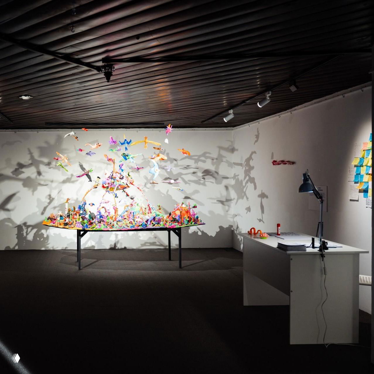 Инсталляция из множества цветных маленьких фигур на столе, драматично отбрасывающая тени на белую стену; рядом рабочий стол с лампой и материалами выставки