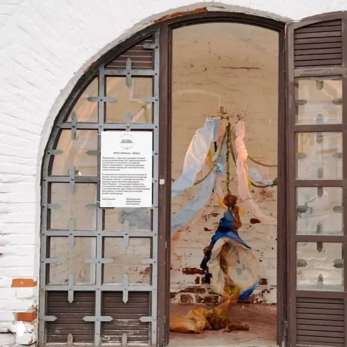 Арочный проём с металлической решёткой, внутри видна произведение искусства на кирпичной стене и информационная табличка у входа в выставочное пространство