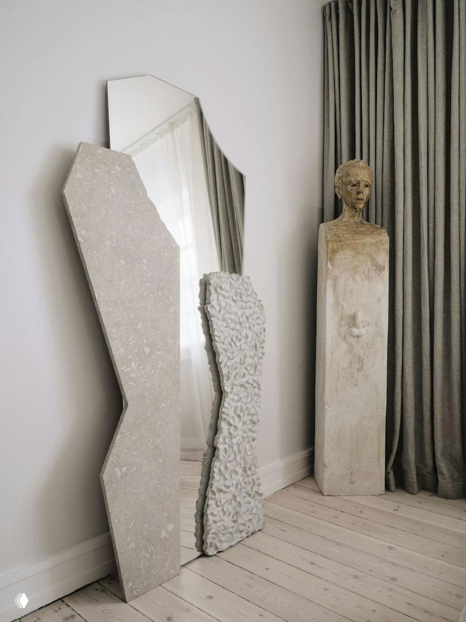 Угол интерьера с крупным фигурным зеркалом, фактурной барельефной панелью и высоким скульптурным бюстом на колонне — минималистичная художественная композиция.