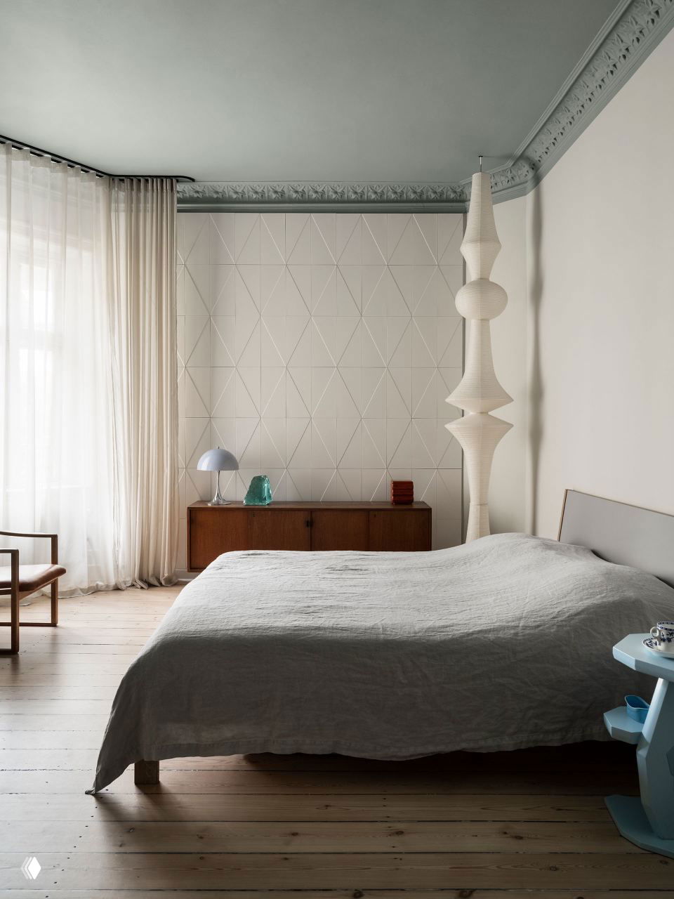 Спальня с пастельными стенами и графичной текстурной панелью за кроватью, высокие окна впускают свет, деревянный комод и мягкое серое постельное бельё.