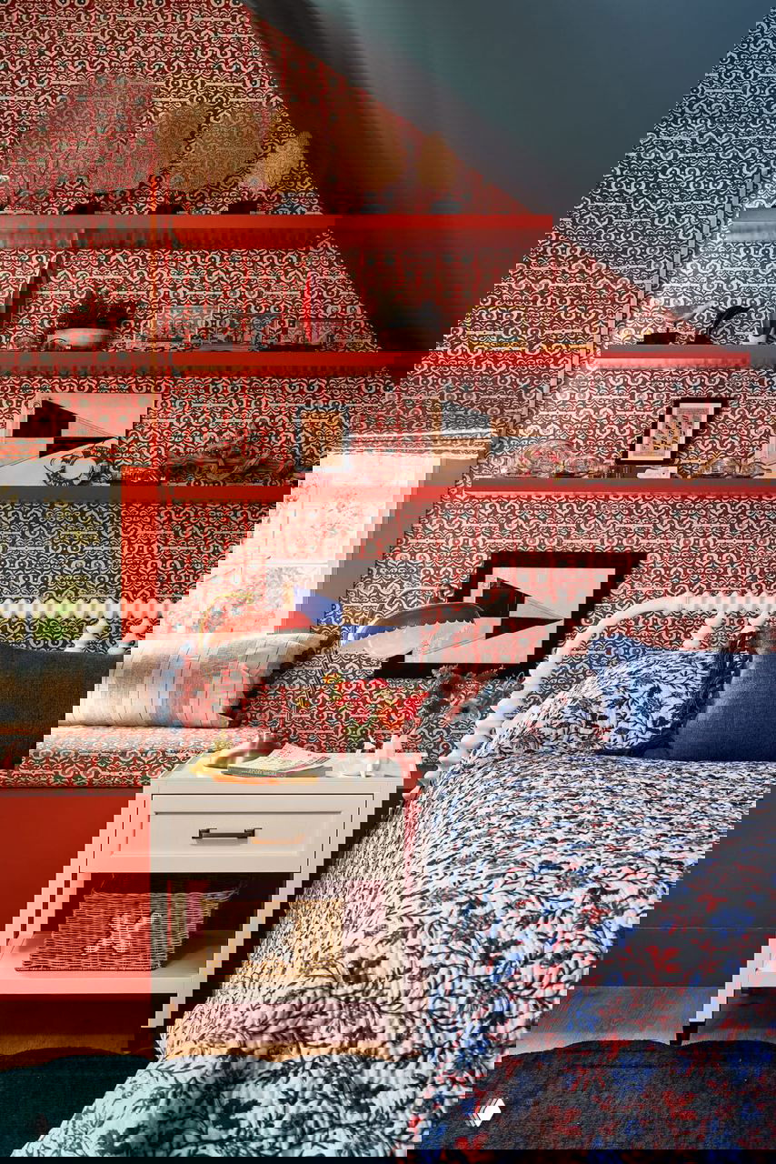 Комната на чердаке с насыщенными красными орнаментальными обоями, открытыми полками и узорным текстилем на кровати и мебели.